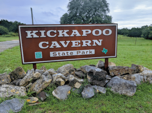 Sign for Kickapoo Cavern State Park, surrounded by rocks and grass, with a cloudy sky in the background.